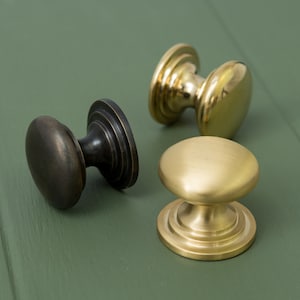 May include: Three cabinet knobs in different finishes: black, brass, and polished brass. The knobs are round with a raised centre and a decorative base.