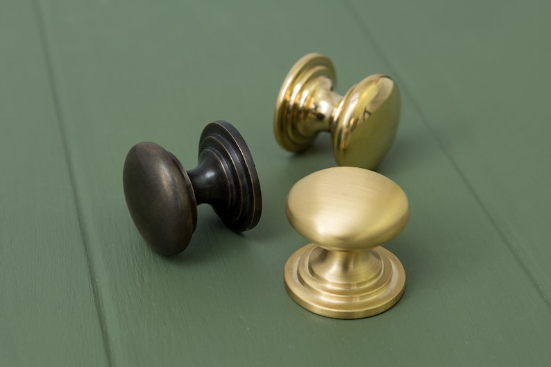 May include: Three cabinet knobs in different finishes: black, brass, and polished brass. The knobs are round with a raised centre and a decorative base.