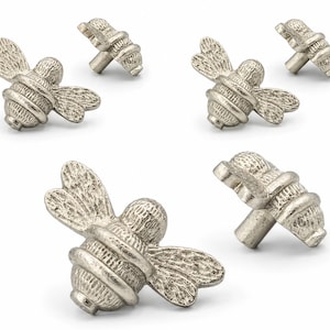 Könnte beinhalten: Ein Set aus sechs silberfarbenen Metall-Möbelknöpfen in Bienenform. Jeder Knopf verfügt über einen strukturierten Körper und Flügel mit metallischem Finish sowie einen zentralen Stift zur Befestigung an Möbelschubladen oder Schranktüren.