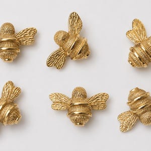May include: Six small, gold-colored metal bee-shaped ornaments. Each bee features a textured body with detailed wings and a smooth, rounded abdomen. The insects are arranged in two rows of three, showcasing a metallic finish and intricate relief.