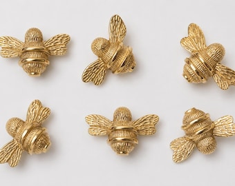 May include: Six small, gold-colored metal bee-shaped ornaments. Each bee features a textured body with detailed wings and a smooth, rounded abdomen. The insects are arranged in two rows of three, showcasing a metallic finish and intricate relief.