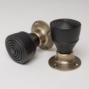 May include: Two black door knobs with a circular design on the handle. The knobs have a brass-colored base with a ridged pattern. The knobs are likely for interior doors and have a classic, vintage style.
