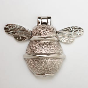 Puede incluir: Un llamador de puerta de metal plateado con forma de abejorro. El insecto presenta un cuerpo redondeado y texturizado con alas detalladas que se extienden hacia los lados y un mecanismo de bisagra liso y pulido en la parte superior.