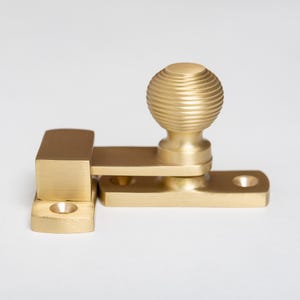 Peut inclure: Une crémone de fenêtre en laiton avec un bouton sphérique décoratif et nervuré. La serrure est composée d'une base rectangulaire, d'un bras pivotant et d'un loquet rectangulaire assorti. La quincaillerie est de couleur dorée chaude.