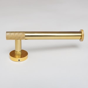 Könnte beinhalten: Ein goldfarbener Toilettenpapierhalter aus Metall mit einer horizontalen Stange in glatter Ausführung sowie einer texturierten Rändelung an der Montagebasis, dem Verbindungspunkt und der Endkappe, zur Wandmontage mit runder Basisplatte.