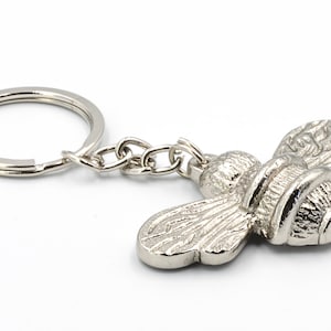 May include: A silver bee keychain with a detailed design. The bee has a round body with wings and a small chain attached to a silver key ring.
