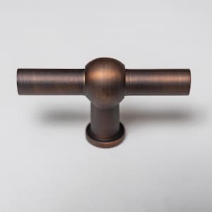 May include: A T-shaped cabinet knob with a dark bronze finish. The hardware features a central spherical hub connecting two horizontal cylindrical bars, supported by a single vertical cylindrical base with a circular mounting plate at the bottom.