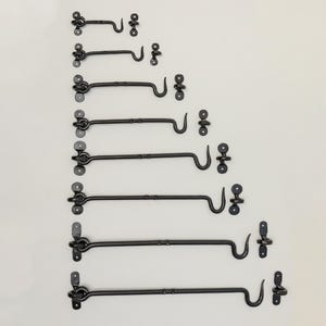 May include: A set of eight black metal hook and eye latches of varying sizes. The latches are arranged in order of size, with the smallest latch at the top and the largest latch at the bottom.