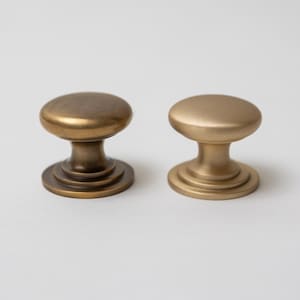 May include: Two round metal cabinet knobs with a smooth, mushroom-shaped top and a stepped circular base. The knob on the left features a dark, antique bronze finish, while the knob on the right has a lighter, brushed satin brass finish.