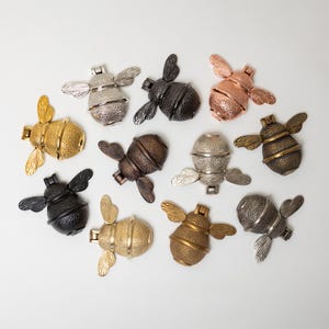 May include: A collection of eleven bee-shaped metal door knockers arranged in a group. Each piece features textured wings and a segmented body, finished in various metallic tones including gold, silver, copper, bronze, black, and gunmetal gray.