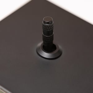 May include: A matte black square plate featuring a central cylindrical knob with a textured, knurled grip pattern. The knob is mounted on a circular base plate, and the entire assembly is finished in a uniform, non-reflective black coating.
