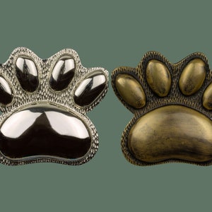 Op de afbeelding: Twee metalen poot afdruk bedels, een zilveren en een gouden. Beide bedels hebben een getextureerd oppervlak en een verhoogd poot afdruk ontwerp.
