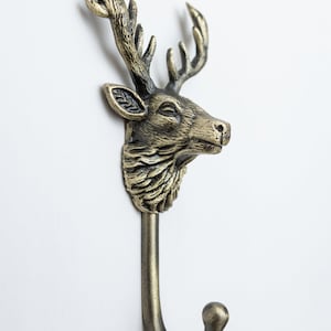 May include: A wall-mounted hook featuring a three-dimensional deer head with detailed antlers and fur texture. The object is finished in an antique brass tone with dark highlights in the crevices, extending into a curved metal hook at the base.