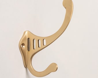 May include: A gold-toned metal wall hook featuring a dual-prong design with a long upper arm and a shorter lower arm. The base plate includes a visible screw hole and four vertical cut-out slots, providing a functional structure for hanging.