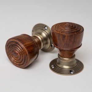 Può includere: Due pomelli antichi per porte. I pomelli sono in legno marrone scuro con un design circolare e sono attaccati a basi in metallo color ottone. Le basi hanno un design circolare con fori per le viti. I pomelli sono su uno sfondo grigio chiaro.