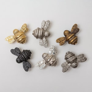 Puede incluir: Seis pomos de metal para armarios con forma de abeja dispuestos en grupo. El conjunto incluye acabados en oro pulido, plata cepillada, bronce antiguo, negro mate, cromo brillante y metal oscuro, cada uno con texturas de alas detalladas.