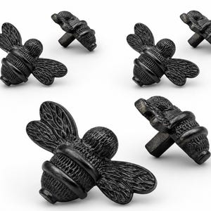 May include: A set of five matte black cabinet knobs shaped like bees. Each knob features detailed wings and a segmented body with a central mounting post on the underside. The bee-shaped hardware is arranged in a group on a plain white background.