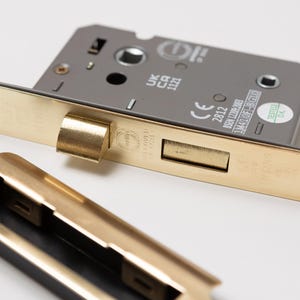 May include: A gold-coloured door lock with a silver faceplate. The lock has a keyhole and a lever handle. The faceplate has the text "UK CA 1121", "CE 2812", "BS EN 12209:2003", "1M4410F-1B-G10", and "TESTED OK".