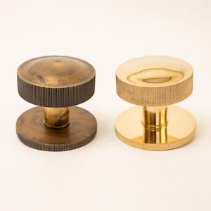 Op de afbeelding: Twee ronde kastknoppen met een cilindrische basis en een platte bovenkant. De ene knop heeft een donkere, verouderde bronzen afwerking met een geribbelde rand, terwijl de andere knop een gepolijste, reflecterende gouden afwerking heeft.
