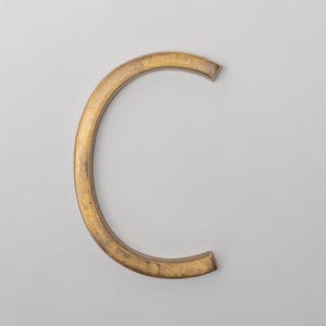May include: A three-dimensional letter C in a metallic gold finish with a textured, weathered surface. The letter has a curved, block-like structure with flat edges and a consistent thickness, presenting a solid, industrial appearance against a plain background.