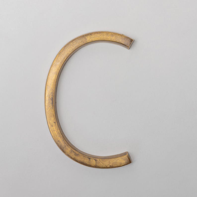 May include: A three-dimensional letter C in a metallic gold finish with a textured, weathered surface. The letter has a curved, block-like structure with flat edges and a consistent thickness, presenting a solid, industrial appearance against a plain background.