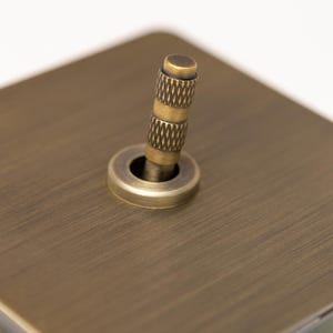 May include: A close-up shot of a vintage light switch. It has a textured, brass-toned toggle and a circular base, set on a brushed metal plate. The design has a classic, industrial feel, reminiscent of older electrical fittings.