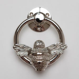 K&ouml;nnte beinhalten: Ein T&uuml;rklopfer aus poliertem, silberfarbenem Metall mit einem kreisf&ouml;rmigen Ring, der an einer runden Montageplatte befestigt ist. Eine detaillierte, texturierte Bienenfigur ist im unteren Teil des Rings zentriert und dient als Klopfer.
