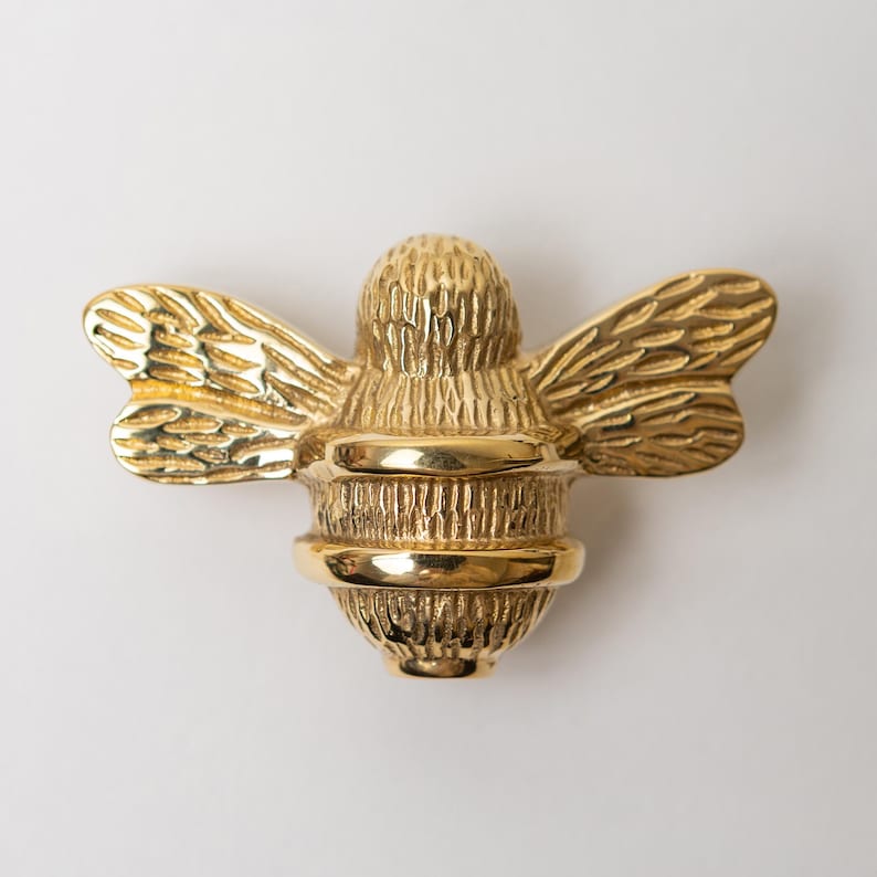 K&ouml;nnte beinhalten: Ein polierter, goldfarbener M&ouml;belknopf in Form einer Biene. Das Insekt verf&uuml;gt &uuml;ber texturierte Fl&uuml;gel und einen segmentierten K&ouml;rper mit einer glatten, reflektierenden Oberfl&auml;che am Mittelband und Kopf, was ein detailliertes, dreidimensionales Aussehen verleiht.