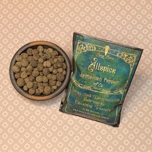 May include: A wooden bowl of allspice berries sits beside a teal and gold packet marked "Allspice Jamaican Pepper." The packet details energy and luck. The background is a pink patterned surface. Measurements are not applicable.