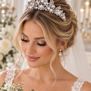 Puede incluir: Una tiara de tono plateado con una disposición simétrica de cristales facetados transparentes y perlas blancas redondas. La pieza presenta un intrincado diseño de metal inspirado en motivos florales, complementado con pendientes de cristal en forma de lágrima.
