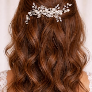 May include: A silver hair comb, embellished with pearls and crystals, sits in wavy auburn hair. The comb has a floral design with pearl clusters and crystal accents, adding elegance. The hair is styled in loose waves, creating a romantic look for special occasions.