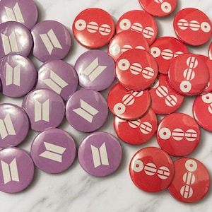 May include: A collection of round buttons in two designs and colors. Some buttons are purple with a white graphic, while others are red with a white graphic. The buttons are scattered across a surface.