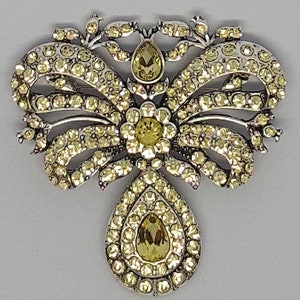 Puede incluir: Un broche plateado con un gran diseño en forma de lágrima. El broche está adornado con numerosas pequeñas piedras brillantes transparentes y presenta una piedra brillante amarilla más grande en el centro.