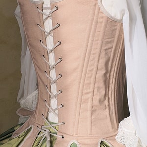 Puede incluir: Un cors&eacute; color melocot&oacute;n con espalda con cordones, que presenta un dise&ntilde;o estructurado con costuras verticales. El cors&eacute; est&aacute; ribeteado con un ribete gris claro y se combina con una blusa blanca y una falda verde. La prenda se exhibe en un maniqu&iacute;.