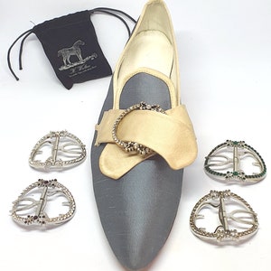 May include: A pair of gray satin shoes with a gold buckle and rhinestones. The shoes are on a white background. There are four additional buckles with rhinestones, two with black stones and two with green stones.
