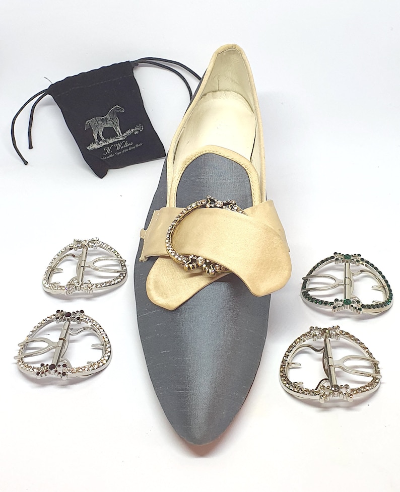 May include: A pair of gray satin shoes with a gold buckle and rhinestones. The shoes are on a white background. There are four additional buckles with rhinestones, two with black stones and two with green stones.
