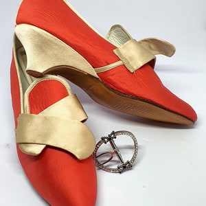 May include: A pair of red satin shoes with gold satin bows and a wedge heel. The shoes are on a white surface with a silver buckle.