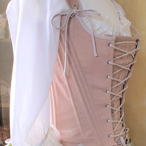 Puede incluir: Un cors&eacute; rosa claro con cordones y correas color crema, combinado con una blusa blanca con ribete de encaje. El cors&eacute; se exhibe en un maniqu&iacute;, mostrando el dise&ntilde;o y la construcci&oacute;n de la prenda hist&oacute;rica.