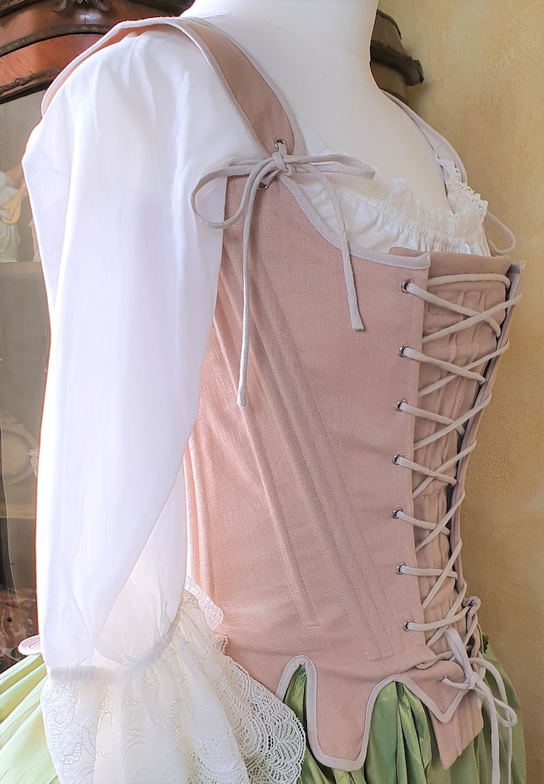 Puede incluir: Un cors&eacute; rosa claro con cordones y correas color crema, combinado con una blusa blanca con ribete de encaje. El cors&eacute; se exhibe en un maniqu&iacute;, mostrando el dise&ntilde;o y la construcci&oacute;n de la prenda hist&oacute;rica.