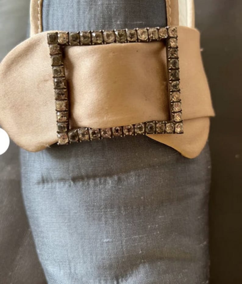May include: A close-up of a beige satin shoe with a large, rectangular buckle adorned with clear rhinestones.