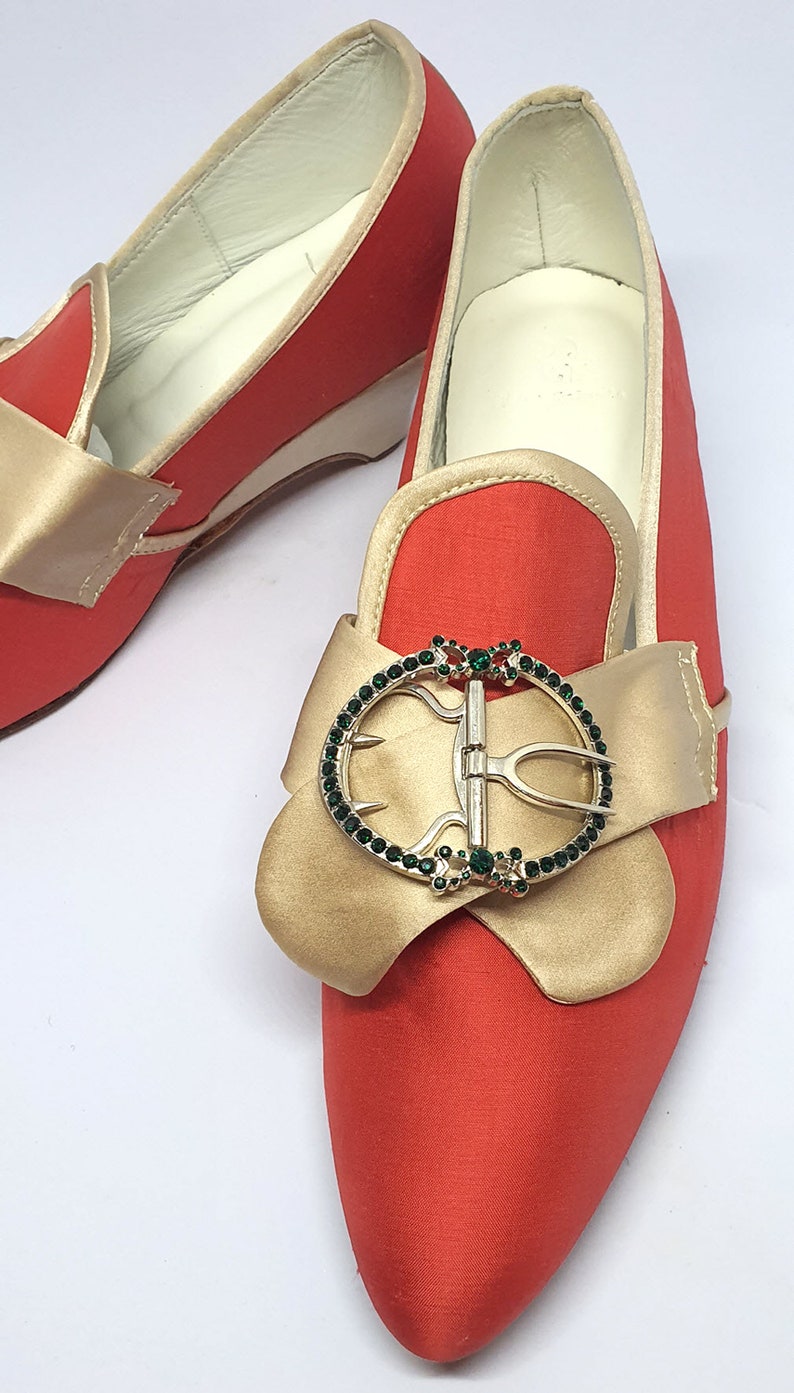 May include: A pair of red satin shoes with gold bows and jeweled buckles. The shoes have pointed toes and a low heel.