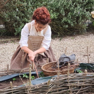 Puede incluir: Una persona con una blusa blanca, una falda marr&oacute;n y un cors&eacute; cuidando un jard&iacute;n. Una valla de mimbre rodea el parterre. Una cesta y un su&eacute;ter gris est&aacute;n cerca. La persona est&aacute; usando una herramienta de madera.