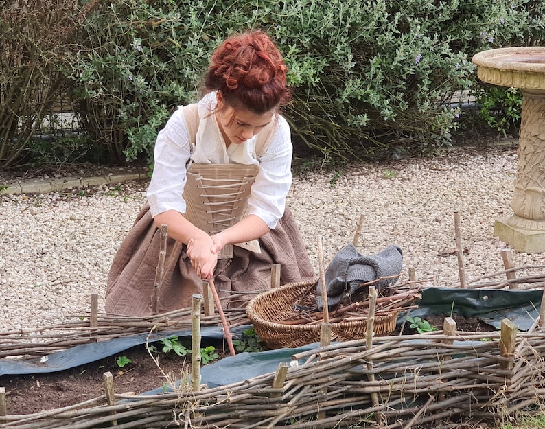 Puede incluir: Una persona con una blusa blanca, una falda marr&oacute;n y un cors&eacute; cuidando un jard&iacute;n. Una valla de mimbre rodea el parterre. Una cesta y un su&eacute;ter gris est&aacute;n cerca. La persona est&aacute; usando una herramienta de madera.