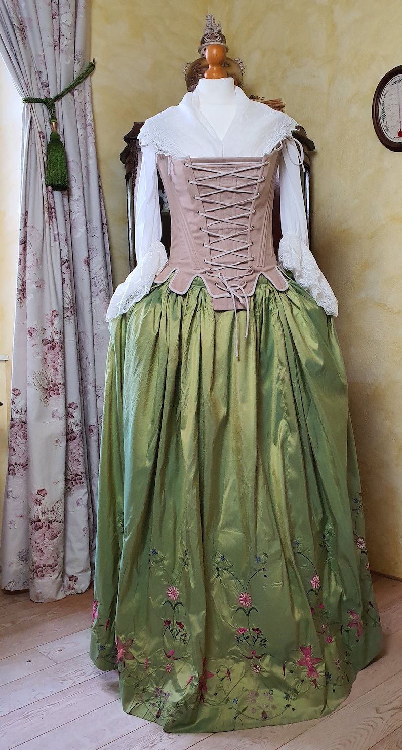 Puede incluir: Un disfraz hist&oacute;rico exhibido en un maniqu&iacute;. El atuendo incluye una blusa blanca con encaje, un cors&eacute; rosa y una falda larga verde con bordados florales. La falda llega hasta el suelo.