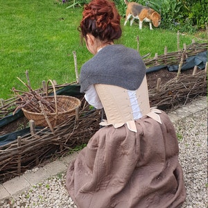 Puede incluir: Una mujer con un chal de punto gris, un cors&eacute; beige y una falda marr&oacute;n estampada. Una cesta de mimbre contiene ramas. Un perro est&aacute; en el fondo sobre un c&eacute;sped verde. La escena es al aire libre.