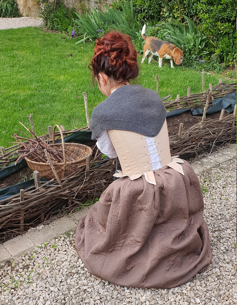 Puede incluir: Una mujer con un chal de punto gris, un cors&eacute; beige y una falda marr&oacute;n estampada. Una cesta de mimbre contiene ramas. Un perro est&aacute; en el fondo sobre un c&eacute;sped verde. La escena es al aire libre.