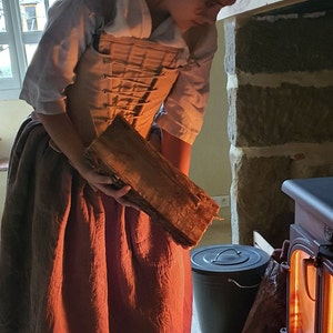 Puede incluir: Una persona con vestimenta hist&oacute;rica sostiene un tronco de madera cerca de una estufa de le&ntilde;a. La persona lleva una blusa blanca, un cors&eacute; beige y una falda larga de color marr&oacute;n rojizo. Un cubo negro est&aacute; cerca.