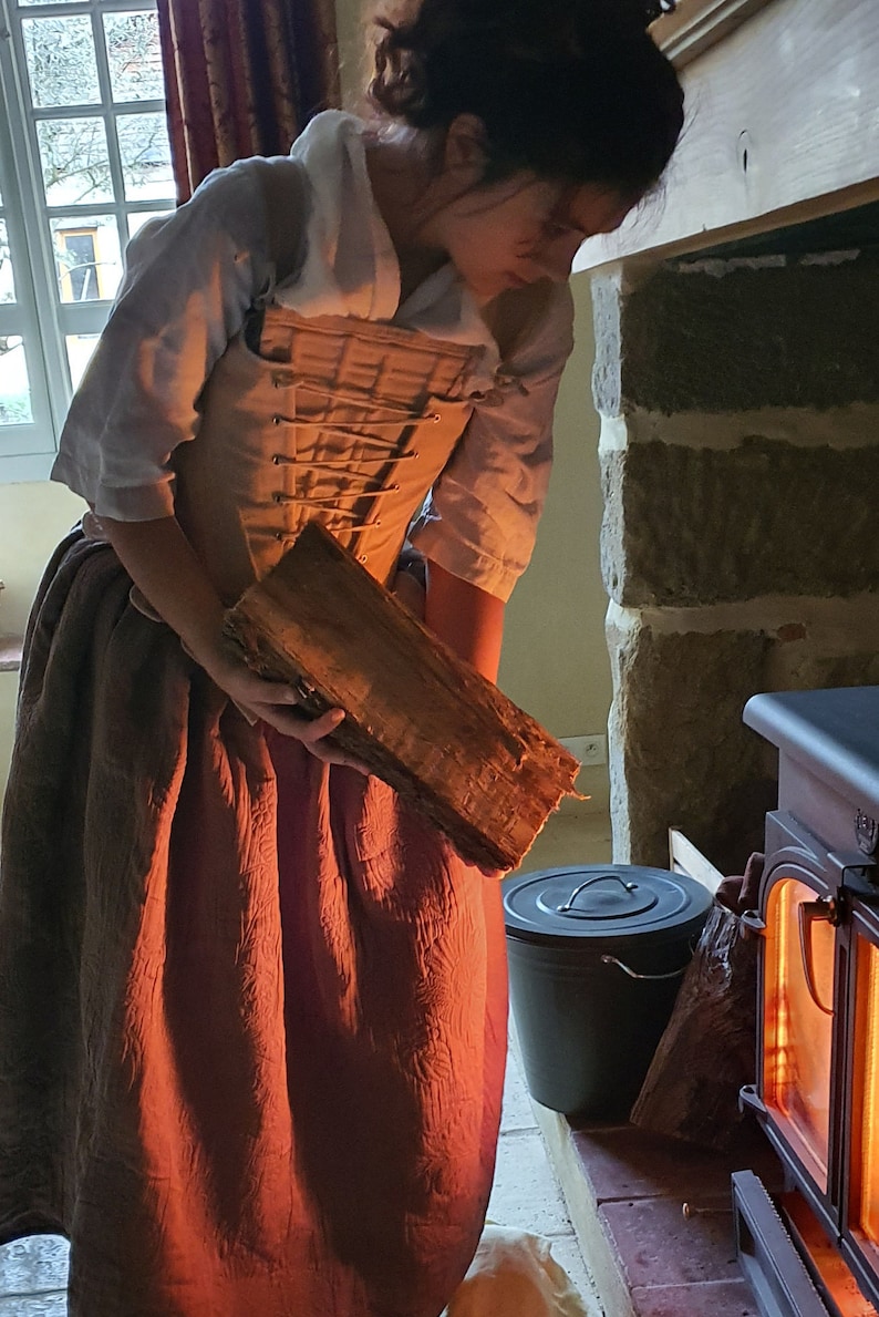 Puede incluir: Una persona con vestimenta hist&oacute;rica sostiene un tronco de madera cerca de una estufa de le&ntilde;a. La persona lleva una blusa blanca, un cors&eacute; beige y una falda larga de color marr&oacute;n rojizo. Un cubo negro est&aacute; cerca.