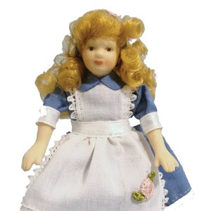 Puede incluir: Una muñeca de porcelana con el pelo rubio rizado que lleva un vestido azul y blanco con ribete de encaje y una rosa rosa bordada en la falda.