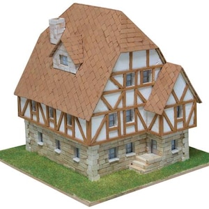 May include: A miniature model of a two-story half-timbered house featuring a brown shingled roof, a stone-textured ground floor, white walls with brown wooden beams, multiple small windows, a chimney, and a set of stone steps at the entrance.
